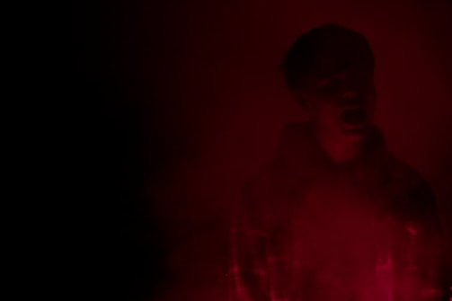 A dark, cinematic close-up of a mysterious figure partially illuminated by subtle red light.