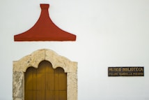 A white wall features a decorative stone archway with a wooden door, topped by a red triangular design. To the right, a black sign with gold text reads 'MUSEO BIBLIOTECA FELIPE CARRILLO PUERTO'.