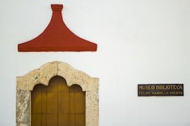 A white wall features a decorative stone archway with a wooden door, topped by a red triangular design. To the right, a black sign with gold text reads 'MUSEO BIBLIOTECA FELIPE CARRILLO PUERTO'.