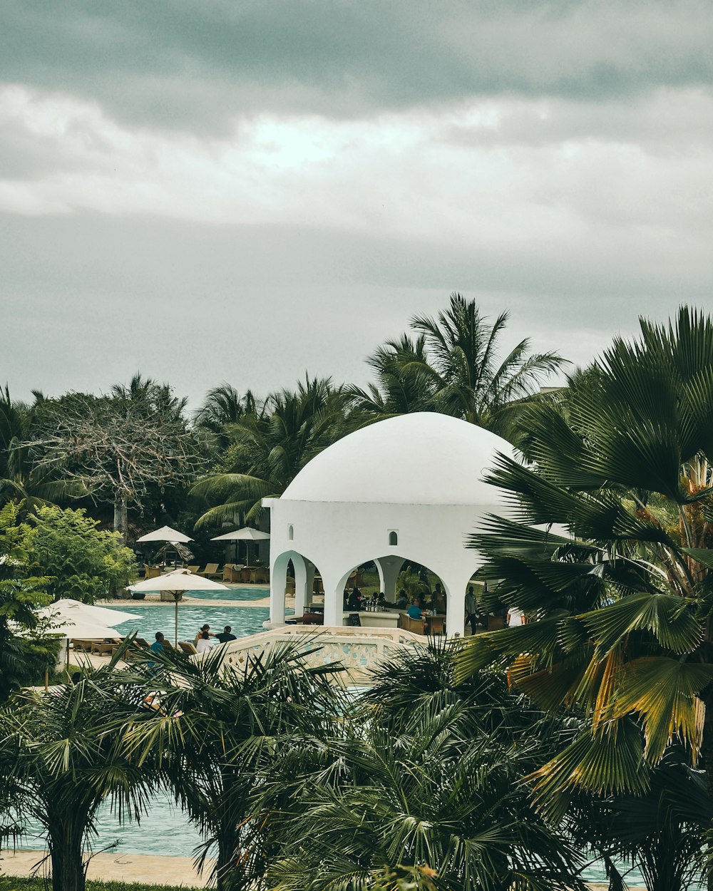 people near swimming pool beside white dome building surrounded with tall and green trees under white and gray skies