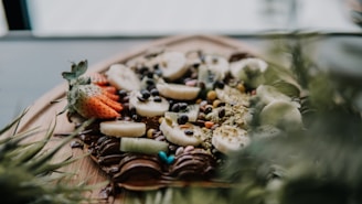 A close-up of a fruit and chocolate dessert topped with sliced bananas, strawberries, kiwis, chocolate chips, and colorful candies. The dessert is placed on a wooden platter, partially surrounded by green leafy plants, creating an inviting and fresh appearance.