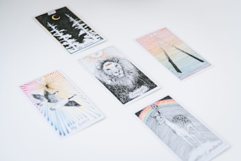 Several tarot cards are spread out on a white surface. Each card features unique illustrations, such as a lion, a crescent moon with forest trees, a pair of flying birds, a deer beneath a rainbow, and two intertwined sticks.
