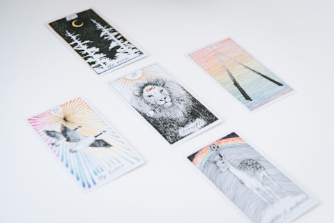 Several tarot cards are spread out on a white surface. Each card features unique illustrations, such as a lion, a crescent moon with forest trees, a pair of flying birds, a deer beneath a rainbow, and two intertwined sticks.
