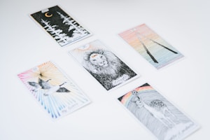 Several tarot cards are spread out on a white surface. Each card features unique illustrations, such as a lion, a crescent moon with forest trees, a pair of flying birds, a deer beneath a rainbow, and two intertwined sticks.