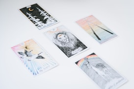 Several tarot cards are spread out on a white surface. Each card features unique illustrations, such as a lion, a crescent moon with forest trees, a pair of flying birds, a deer beneath a rainbow, and two intertwined sticks.
