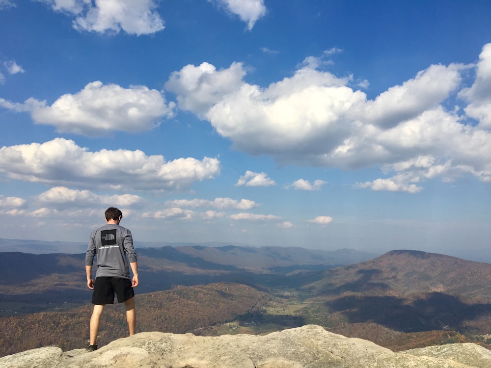Mann im grauen langärmeligen Hemd steht tagsüber am Rand der Klippe über Berge unter bewölktem Himmel