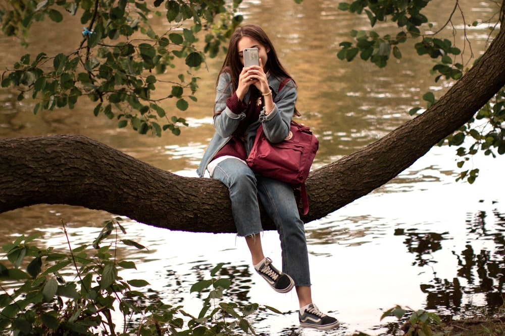 girl sitting on tree branch near the body of water during daytime ]