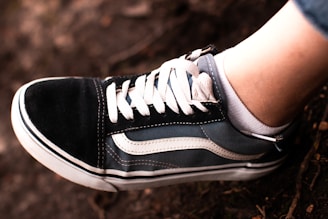 A close-up of a black and white sneaker with a distinctive wave-like pattern on the side, laced with white shoelaces. The shoe is being worn by a person, whose ankle and a portion of their denim jeans are visible.