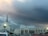 A large church building with a prominent steeple topped with a cross is situated amidst a parking lot filled with cars. Overhead, the sky is dramatic with dark storm clouds gathering, yet a subtle hint of sunlight penetrates, casting a slight glow.