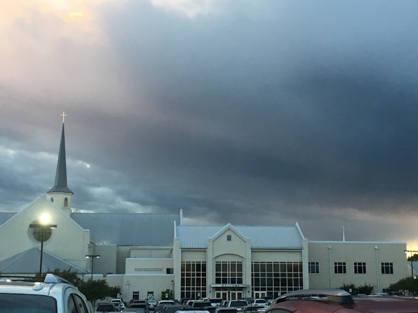 A large church building with a prominent steeple topped with a cross is situated amidst a parking lot filled with cars. Overhead, the sky is dramatic with dark storm clouds gathering, yet a subtle hint of sunlight penetrates, casting a slight glow.