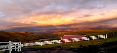 A picturesque rural landscape features rolling hills under a dramatic sky with warm, golden light. A red barn stands on the grassy hill, enclosed by white fencing that accentuates the scene’s tranquil and bucolic atmosphere.