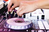 A person is adjusting controls on a DJ mixer, with a focus on their hands and the equipment. The mixer features various dials, buttons, and a jog wheel, suggesting a dynamic and engaging setting.