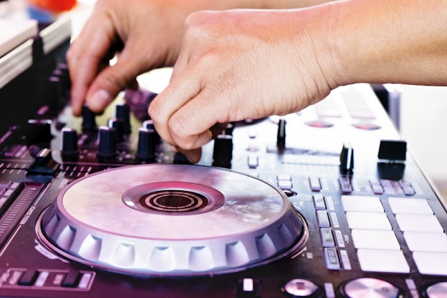 A person is adjusting controls on a DJ mixer, with a focus on their hands and the equipment. The mixer features various dials, buttons, and a jog wheel, suggesting a dynamic and engaging setting.