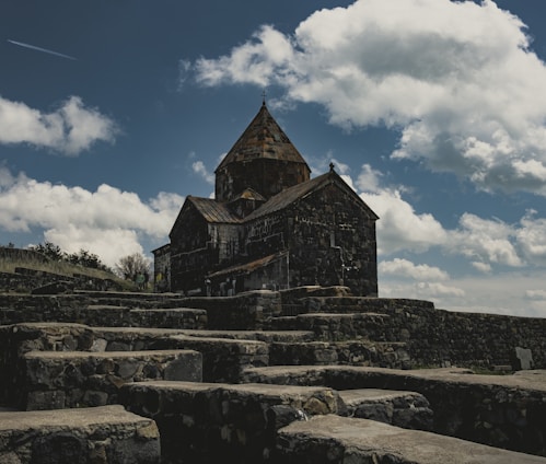A historic stone monastery is situated atop a stepped stone platform under a partly cloudy sky. The architecture features an intricate design with a prominent dome, creating a sense of ancient serenity.