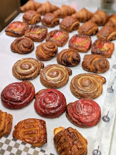 A variety of baked pastries are displayed on a tray. There are swirled, circular pastries with glossy surfaces in red and brown hues. Rectangular pastries with a topping that appears to be rhubarb are visible, as well as several croissant-like pastries with flaky, golden brown crusts.