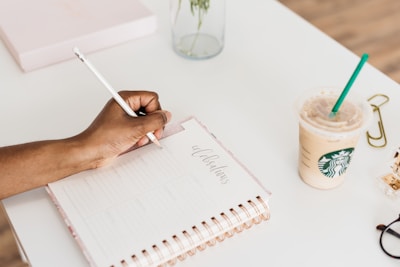 A person writing in a planner with a white pen on a clean white table. The planner is open to a page labeled 'celebrations.' Nearby, there is a Starbucks iced coffee with a green straw, gold paperclips, and a transparent tape dispenser. A glass vase with a plant is also partially visible.