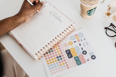 A person is writing in a planner notebook with a copper spiral binding, surrounded by colorful stickers. There is a beige-colored drink with a green logo in a cup nearby, alongside a pair of glasses and a stapler on a white table.