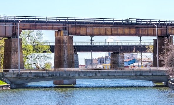 Three bridges span a body of water. The foremost bridge is concrete with metal railings, while behind it, two more bridges made of rusted metal and concrete support structures cross over the same waterway. Trees with fresh green foliage and a structure resembling an industrial building are visible in the background.