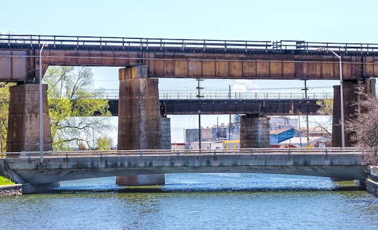 Three bridges span a body of water. The foremost bridge is concrete with metal railings, while behind it, two more bridges made of rusted metal and concrete support structures cross over the same waterway. Trees with fresh green foliage and a structure resembling an industrial building are visible in the background.