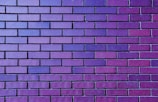 A wall composed of uniformly sized rectangular bricks arranged in a horizontal pattern. The bricks display a gradient of colors ranging from deep purple to a lighter lavender, creating a rich visual texture. The mortar lines are consistently spaced, emphasizing the orderly design.