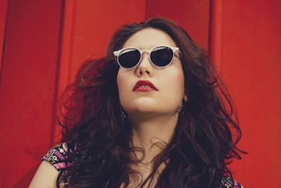 A person with long curly hair wearing white-framed sunglasses leans against a vibrant red background. Their head is tilted back slightly, and they have a confident expression with bold red lipstick matching the backdrop. The clothing features a colorful pattern, adding to the stylish and bold appearance.