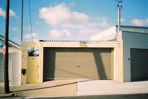 A garage door and the entrance of a suburban house with a yellow gate marked with the number 23. The building has a flat facade and a slightly industrial look with concrete structures. The scene is lit by sunlight, casting shadows on the garage door.