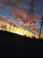 A silhouette of high-voltage power lines and pylons against a vibrant sunset sky. The sky displays a gradient of deep blue, orange, and yellow hues as the sun sets on the horizon. Sparse trees and urban elements are visible at the base of the power structures.