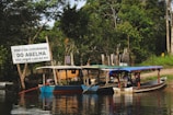 A serene riverside scene featuring several wooden boats moored along a lush, green shoreline. A noticeable sign reads 'Porto da Comunidade do Abelha' with a message asking not to throw waste into the river. The area is surrounded by dense vegetation and trees, with a rustic fence visible in the background.