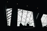 The image features a modern architectural interior with a prominent glass ceiling structured with intersecting metal beams. Silhouettes of people can be seen walking near the railing, which adds depth and scale to the scene.