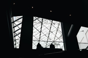 The image features a modern architectural interior with a prominent glass ceiling structured with intersecting metal beams. Silhouettes of people can be seen walking near the railing, which adds depth and scale to the scene.