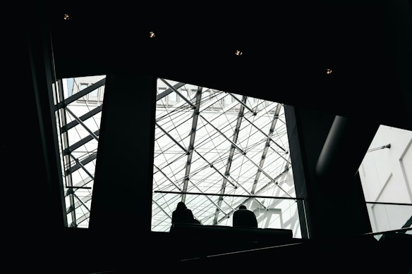 The image features a modern architectural interior with a prominent glass ceiling structured with intersecting metal beams. Silhouettes of people can be seen walking near the railing, which adds depth and scale to the scene.