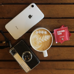 A white iPhone is placed on a wooden table alongside a vintage camera and a cup of cappuccino with latte art. Two sugar packets labeled 'Coffee Day' are positioned nearby.