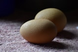 Two brown eggs are placed on a textured surface, with droplets of water visible on their shells. The lighting is soft and moody, casting gentle shadows around the eggs.