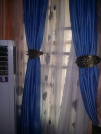 A pair of blue curtains with intricate golden tiebacks is partially covering a window. Behind them, sheer curtains featuring a subtle pattern allow light to filter through. An air conditioning unit is visible on the left side of the image.