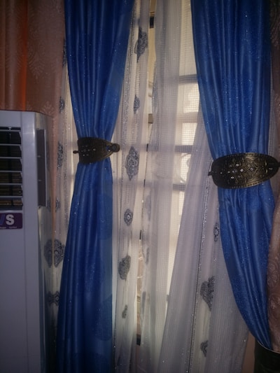 A pair of blue curtains with intricate golden tiebacks is partially covering a window. Behind them, sheer curtains featuring a subtle pattern allow light to filter through. An air conditioning unit is visible on the left side of the image.