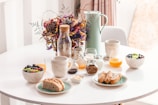 A round table is set with a variety of breakfast items, including bread, berries, a bowl of granola, jars of honey and milk, and two cups of coffee. A pitcher and a glass of orange juice accompany the spread. In the center, a decorative bouquet of dried flowers adds an aesthetic touch.
