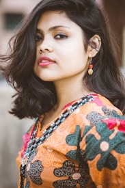 A person with medium-length black hair is wearing vibrant, patterned clothing with floral designs. The clothing features intricate beadwork and bright colors, including orange, black, and red. The person is accessorized with a dangling earring that includes a bead and a shell-like pendant.