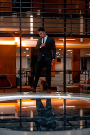 A man in a suit stands confidently indoors with a modern architectural background. The warm lighting creates a dramatic reflection in a glassy surface below, enhancing the visual symmetry.
