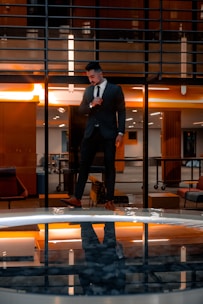 A man in a suit stands confidently indoors with a modern architectural background. The warm lighting creates a dramatic reflection in a glassy surface below, enhancing the visual symmetry.