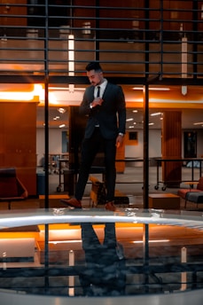 A man in a suit stands confidently indoors with a modern architectural background. The warm lighting creates a dramatic reflection in a glassy surface below, enhancing the visual symmetry.