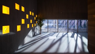 An interior space with a dark wall featuring illuminated yellow squares, each containing an abstract design. A potted plant is placed near a wall with large floor-to-ceiling windows that allow natural light to cast long shadows on the floor.