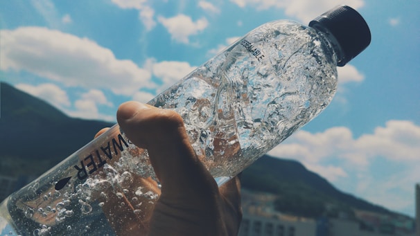 A hand holding a clear plastic water bottle, filled with water, against a backdrop of a blue sky with white clouds. The light shines through the bottle, creating a glistening effect on the water inside. The silhouette of a mountain range is visible in the background.