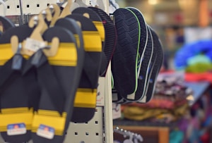 A store display features multiple pairs of flip-flops hanging on hooks. The foreground shows black and yellow flip-flops, while the background reveals several pairs with darker soles and green accents. The surrounding environment is slightly blurred, indicating a retail setting with various colorful fabrics visible in the distance.