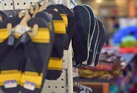 A store display features multiple pairs of flip-flops hanging on hooks. The foreground shows black and yellow flip-flops, while the background reveals several pairs with darker soles and green accents. The surrounding environment is slightly blurred, indicating a retail setting with various colorful fabrics visible in the distance.