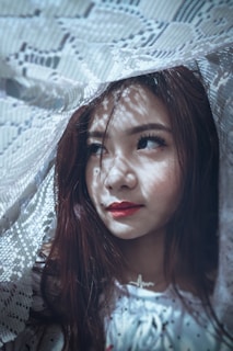 A woman is portrayed with a pensive expression, surrounded by delicate, intricate lace creating ornate shadows on her face. Her makeup is subtle but elegant, accentuated by a bold red lip.