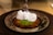 A close-up of a dessert featuring a crispy, golden pastry topped with a dollop of fluffy white cream and a sprinkle of finely chopped green pistachios. The dessert is presented on a round metal plate, and a fork is poised above it.
