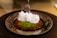 A close-up of a dessert featuring a crispy, golden pastry topped with a dollop of fluffy white cream and a sprinkle of finely chopped green pistachios. The dessert is presented on a round metal plate, and a fork is poised above it.