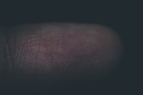 A close-up view of a human fingertip showing detailed fingerprint ridges and grooves, with minimal lighting highlighting the texture of the skin.