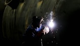 A person wearing protective gear, including a welding mask, is engaged in welding work on a large metallic surface. Bright sparks and a glow are emitted from the welding point, contrasting against the dark environment.