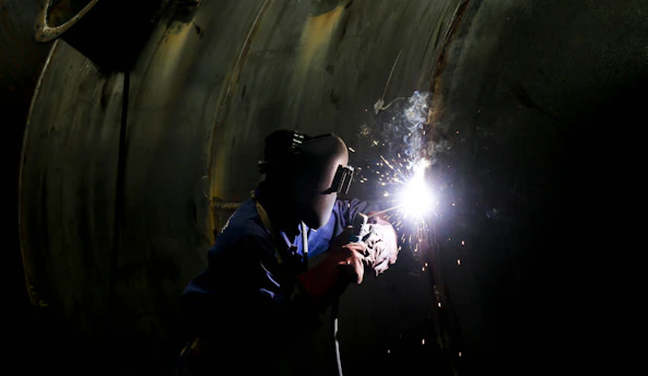 A person wearing protective gear, including a welding mask, is engaged in welding work on a large metallic surface. Bright sparks and a glow are emitted from the welding point, contrasting against the dark environment.